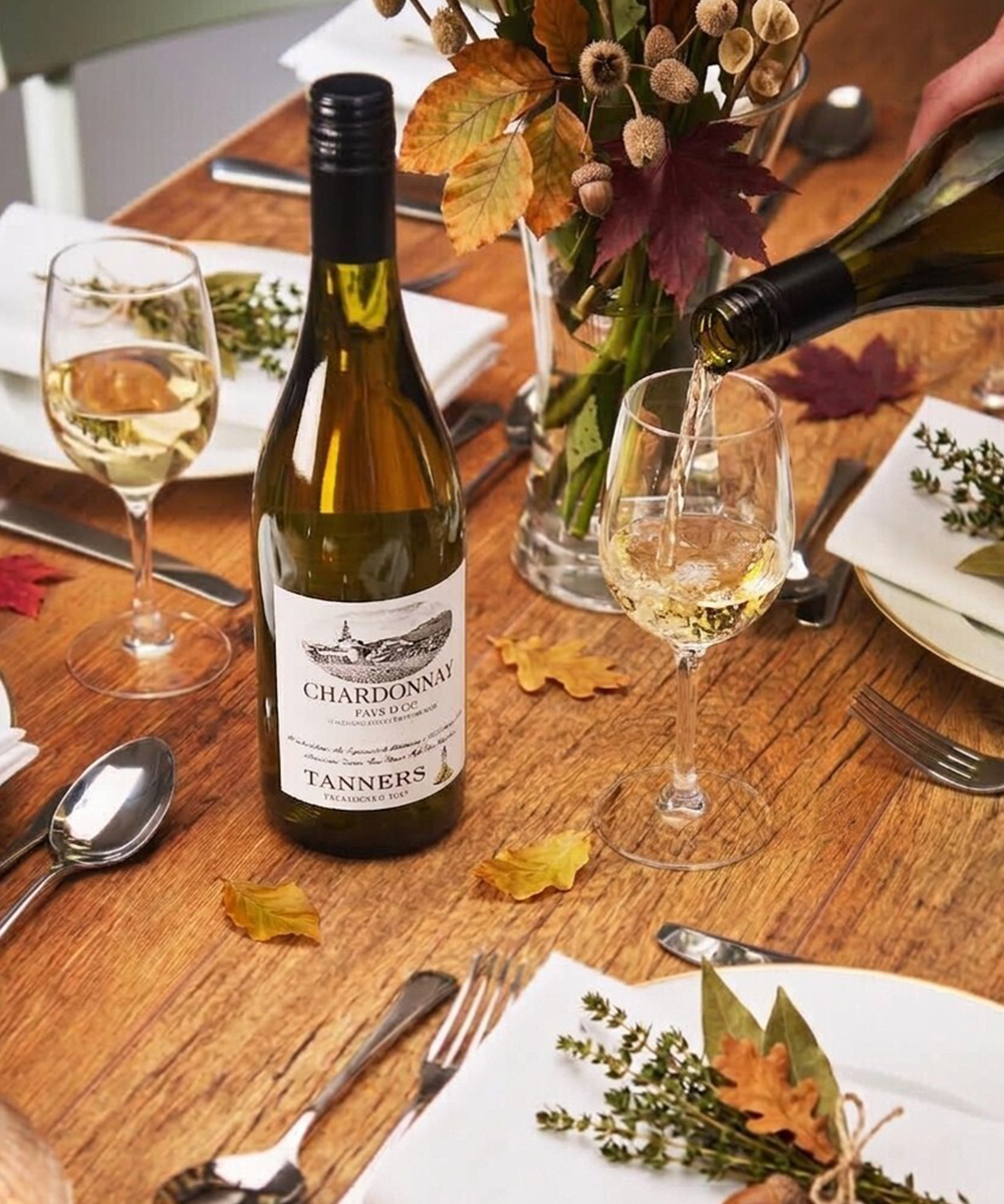 A bottle of Chardonnay from Tanners is shown on a rustic wooden dining table, with a hand pouring wine from a second bottle into a glass. The table is set with white plates, silver cutlery, and autumn-themed floral arrangements.