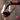 Red wine being poured from a dark bottle with a red foil capsule into a clear stemmed glass, set against a blurred stone-textured background. A red circular logo watermark is visible in the bottom right corner.