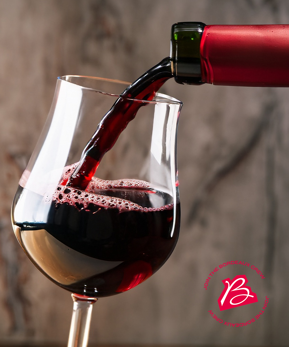 Red wine being poured from a dark bottle with a red foil capsule into a clear stemmed glass, set against a blurred stone-textured background. A red circular logo watermark is visible in the bottom right corner.