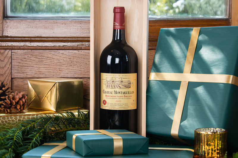 A bottle of Château Montaiguillon red wine in a light wood presentation box, styled with teal and gold gift boxes, pinecones, and evergreen branches in front of a rustic wooden window.