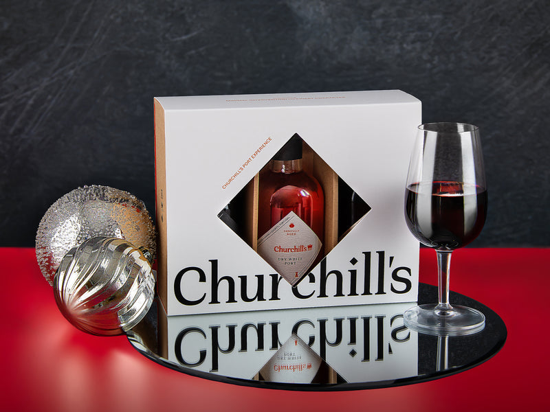 Churchill's port gift set with a 3 bottles in gift box, and glass on a reflective surface.