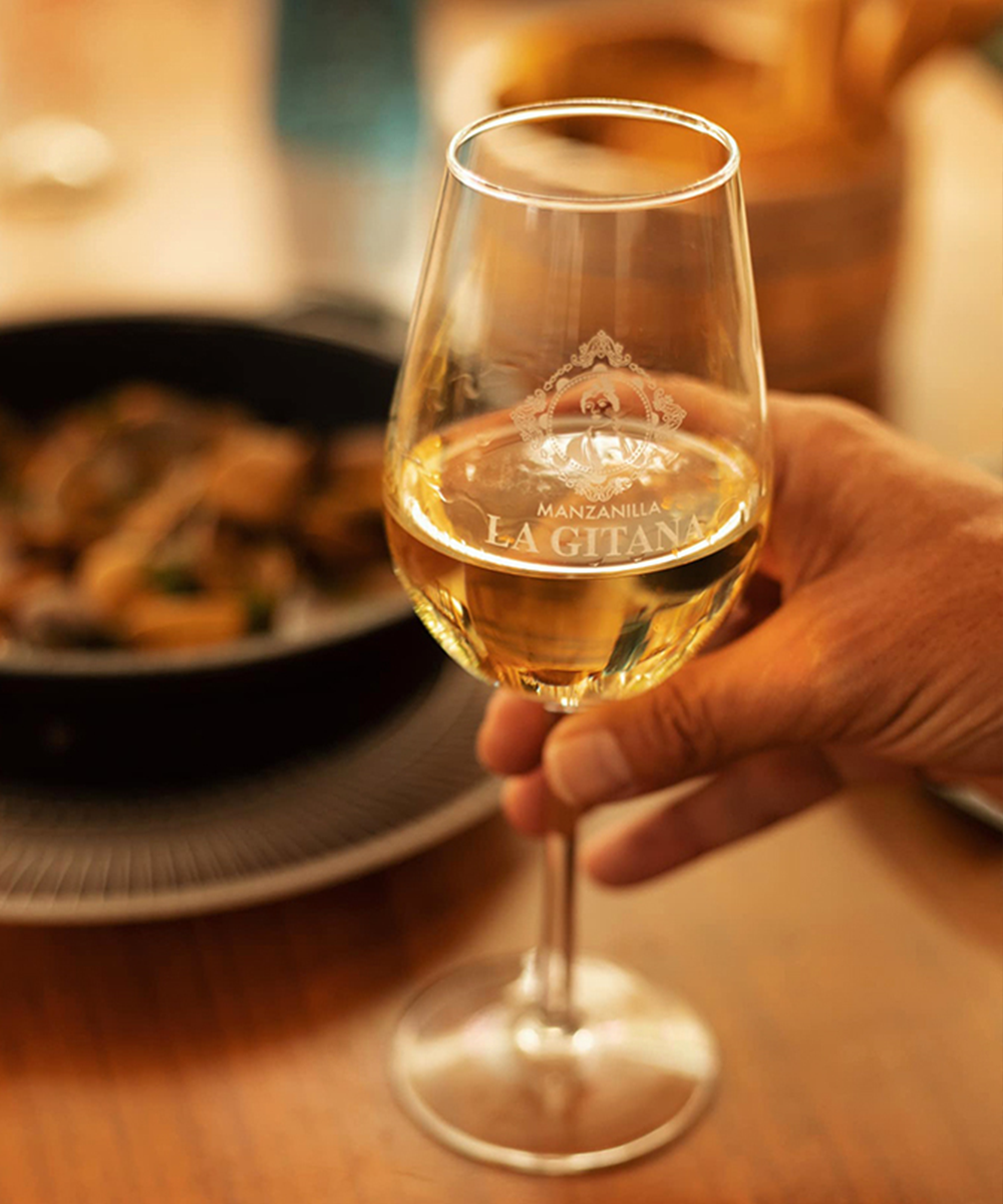 Glass of white wine with 'La Manzanilla' branding held by a hand, blurred background