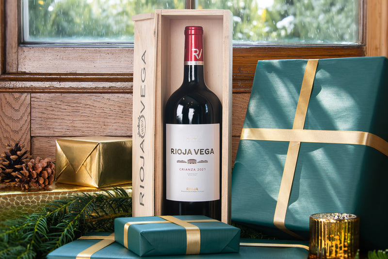 A bottle of Rioja Vega red wine in a light wood presentation box, styled with teal and gold gift boxes, pinecones, and evergreen branches in front of a rustic wooden window.