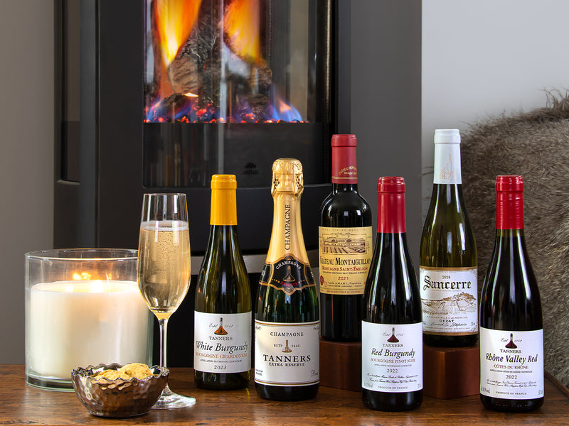 Assorted wine bottles on a wooden table with a candle and glass in front of a fireplace.