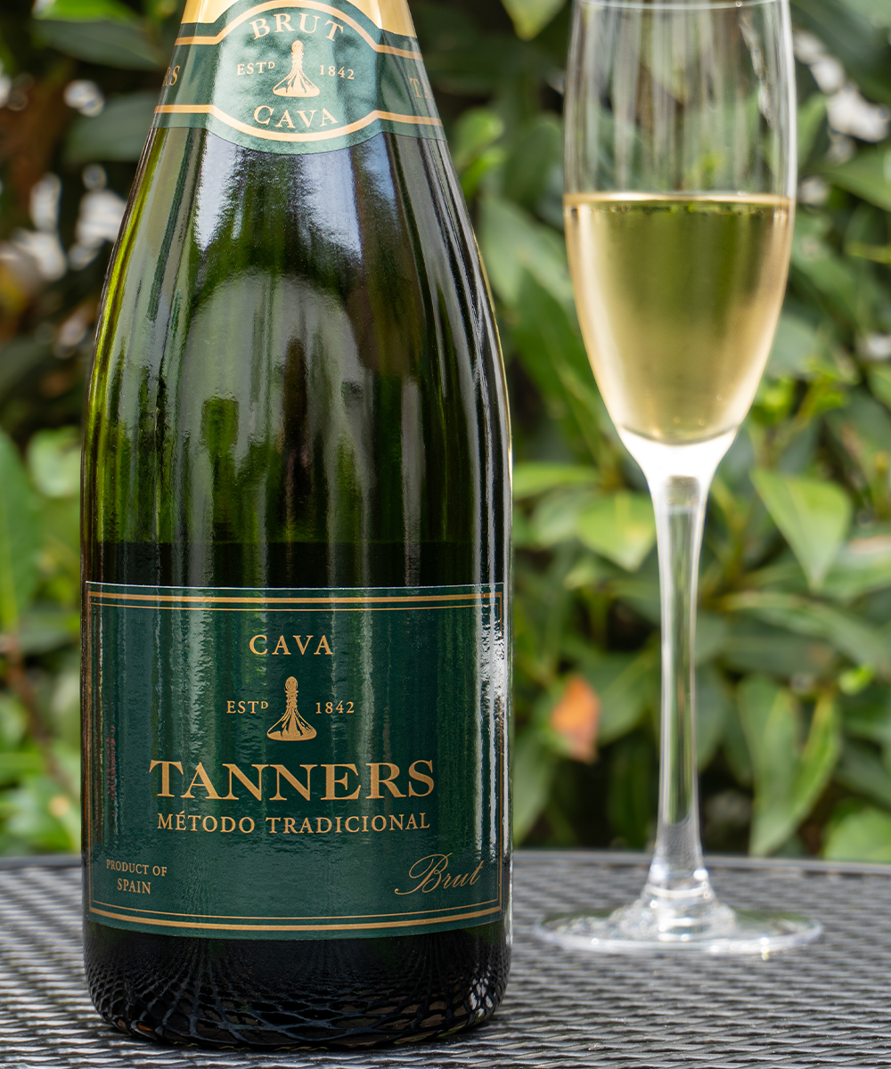 A bottle of Tanners Cava Brut Espumante with a dark green label and gold lettering, standing next to a filled champagne flute on a black mesh table with a blurred green garden background.
