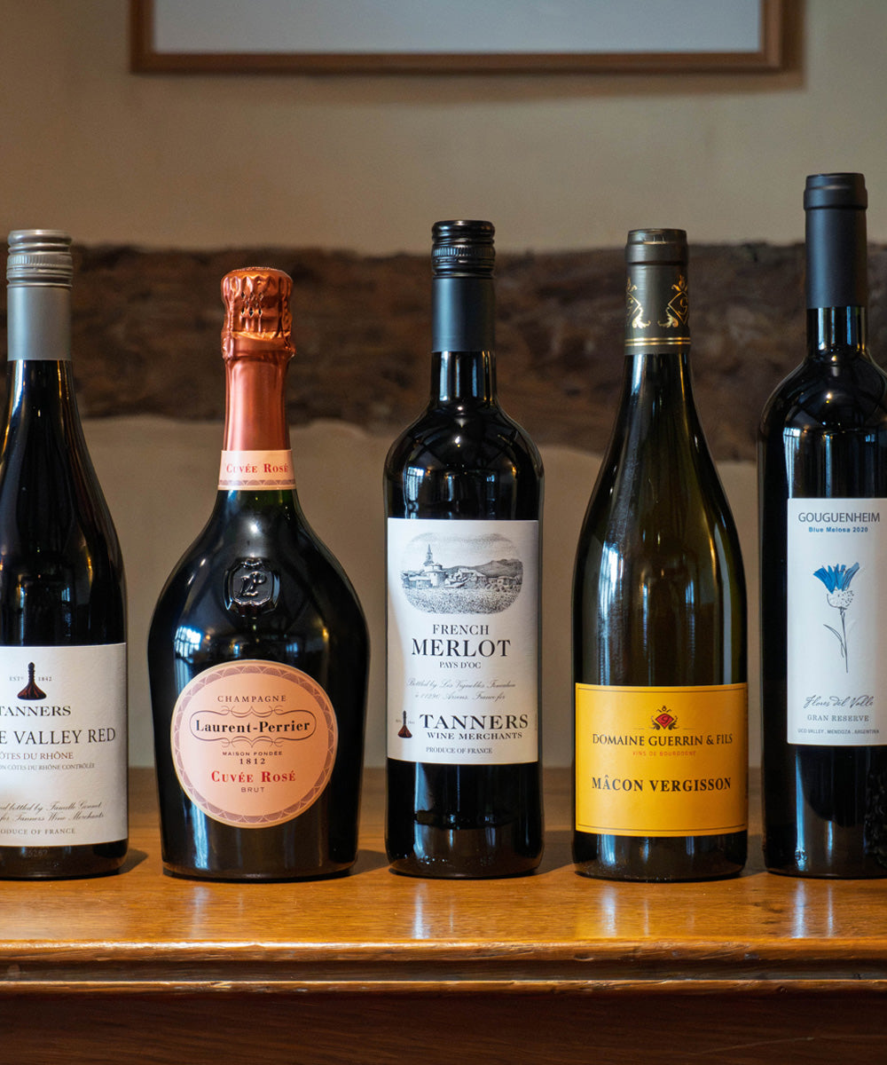 Five wine bottles lined up on a wooden shelf in front of a rustic stone wall. The bottles include wines from Tanners, Laurent Perrier, and Domaine Guerrin & Fils.