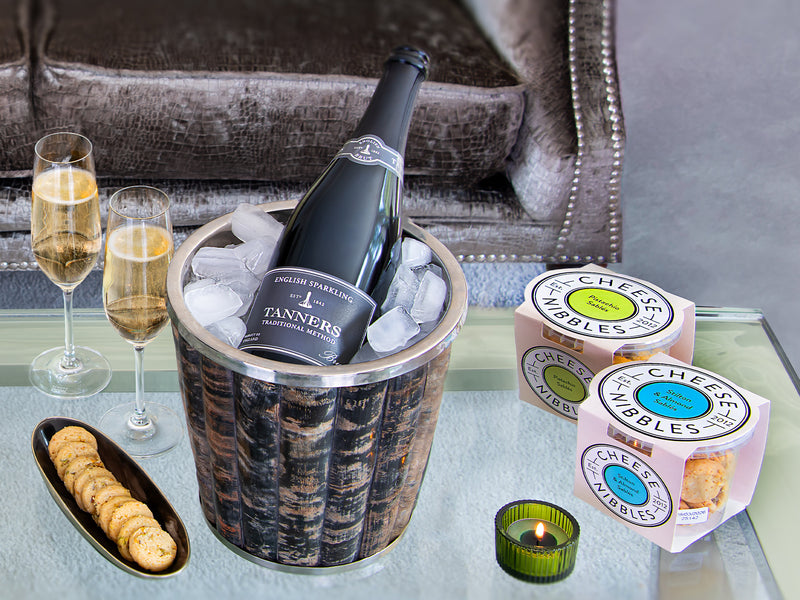 Bottle of Tanners champagne in an ice bucket with two glasses, a tray of nibbles, and boxes of Cheese Nibbles on a table.