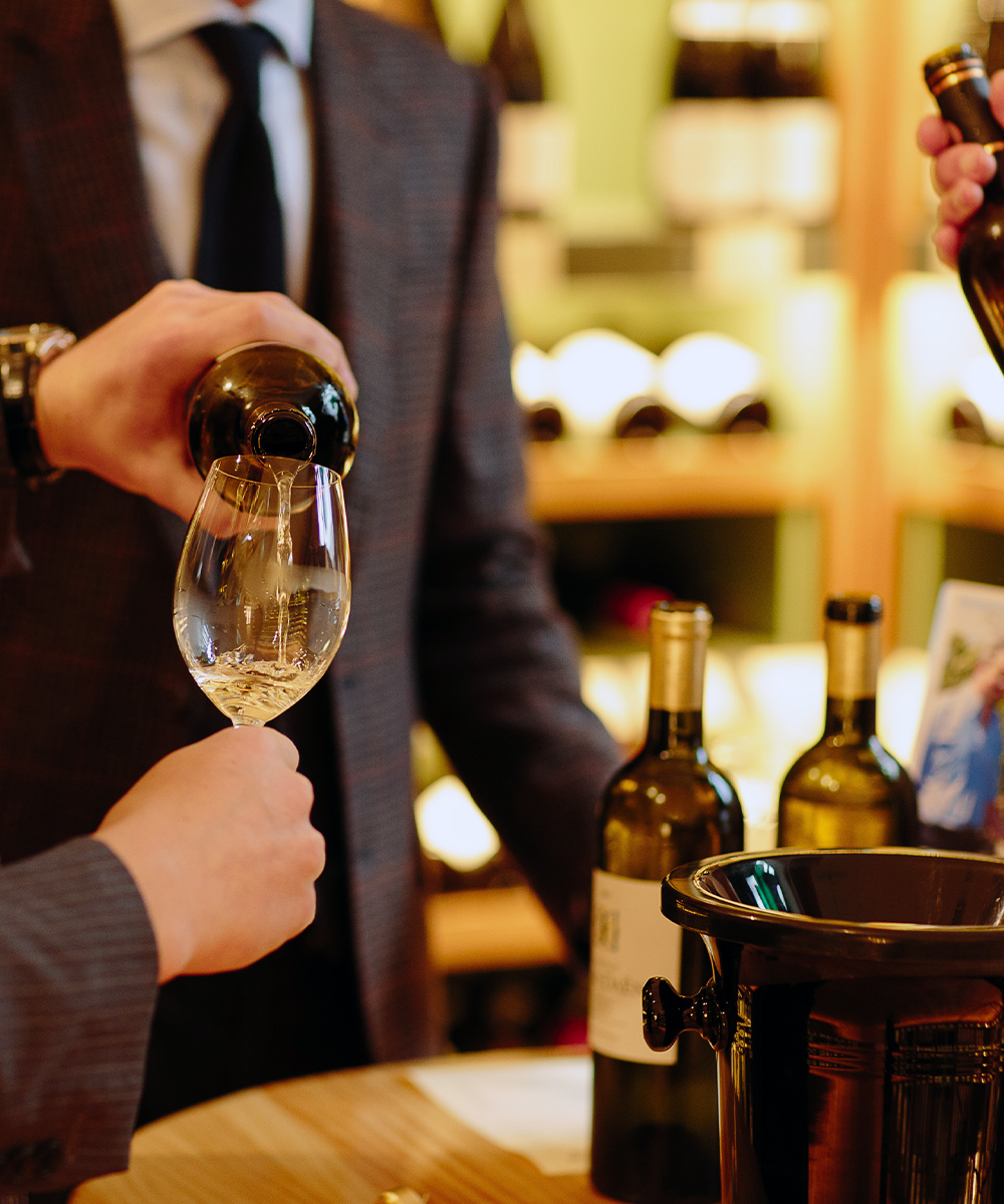 Person pouring white wine into a glass with bottles and a wine cooler in the background.