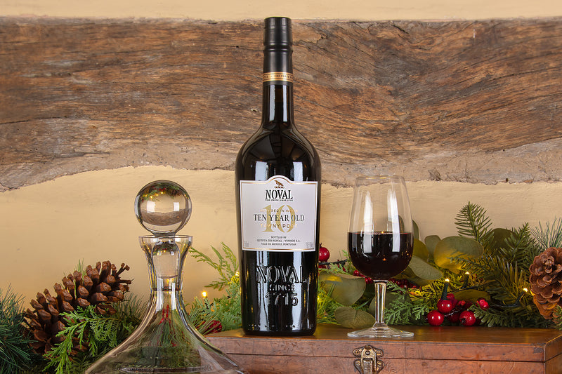 A bottle of Noval Ten Year Tawny Port stands on a wooden surface beside a filled wine glass, a clear glass decanter, and holiday greenery with pinecones and red berries, set against a rustic wood and beige wall background.