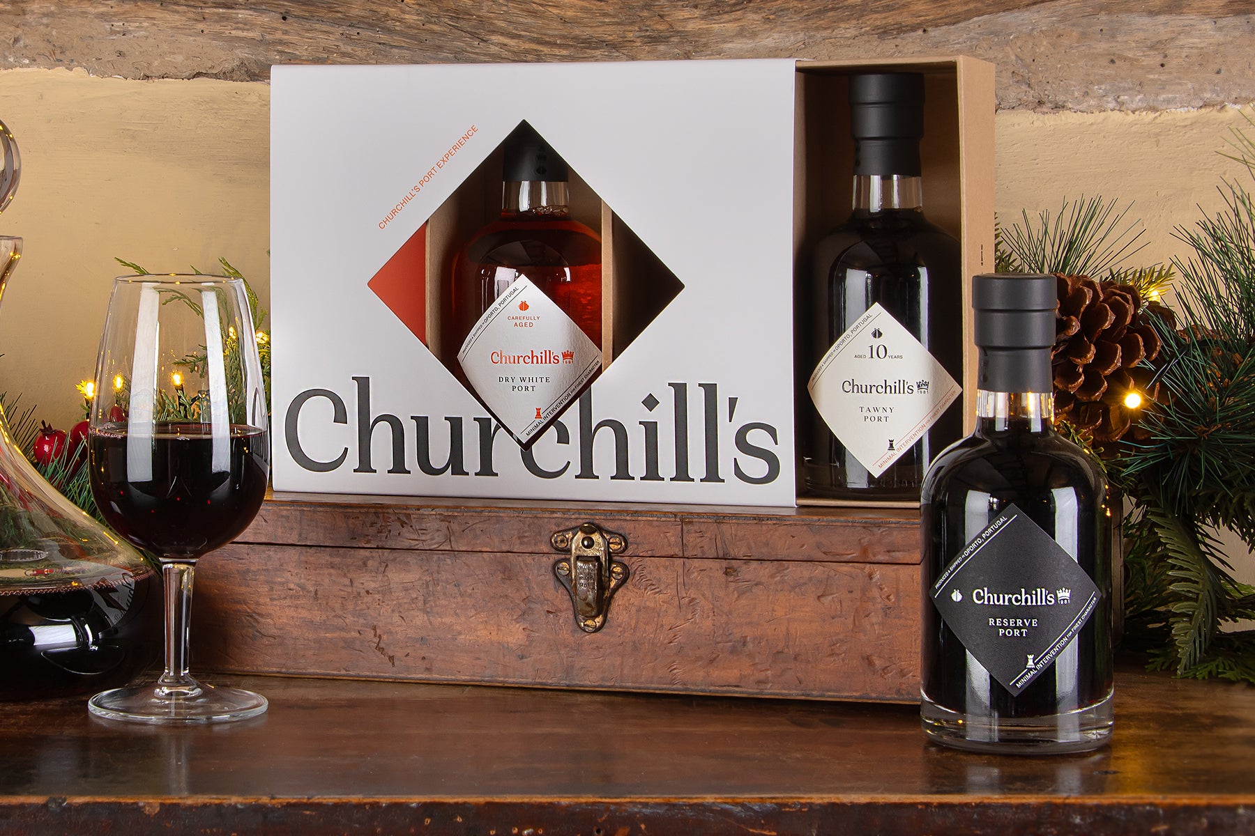 Churchill's Port experience gift set, featuring a white, ruby and tawny port presented on a wooden box with a white front panel, two bottles of port inside, and a third bottle placed in front on a wooden table, with a glass of port and festive greenery in the background.