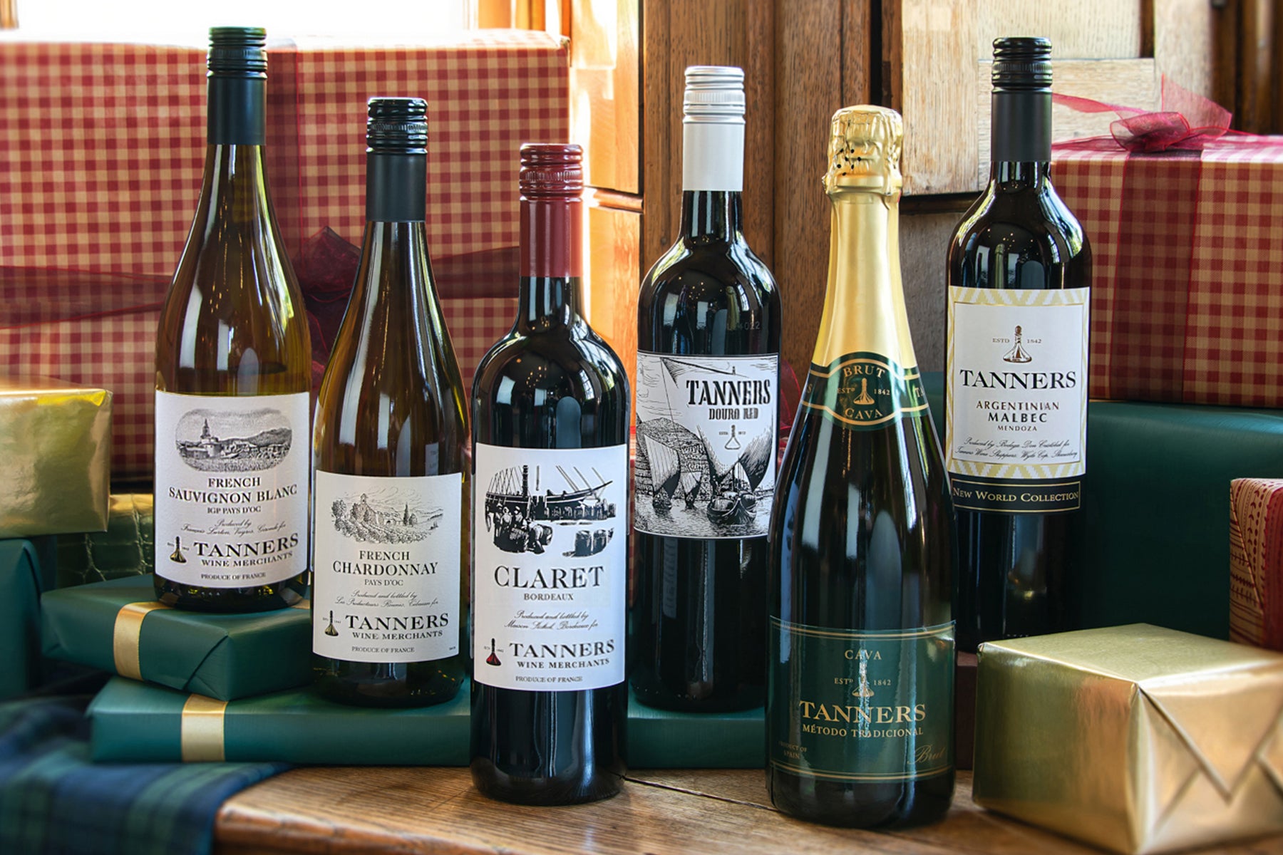 A collection of Tanners wine bottles including a bottle of French Sauvignon Blanc, French Chardonnay, Claret Bordeaux, Douro Red and Argentinian Malbec alongside a bottle of Corona Gold champagne arranged on a wooden table with wrapped gift boxes in the background.