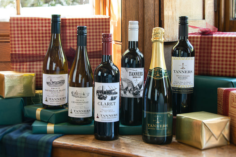 A collection of Tanners wine bottles including a bottle of French Sauvignon Blanc, French Chardonnay, Claret Bordeaux, Douro Red and Argentinian Malbec alongside a bottle of Corona Gold champagne arranged on a wooden table with wrapped gift boxes in the background.