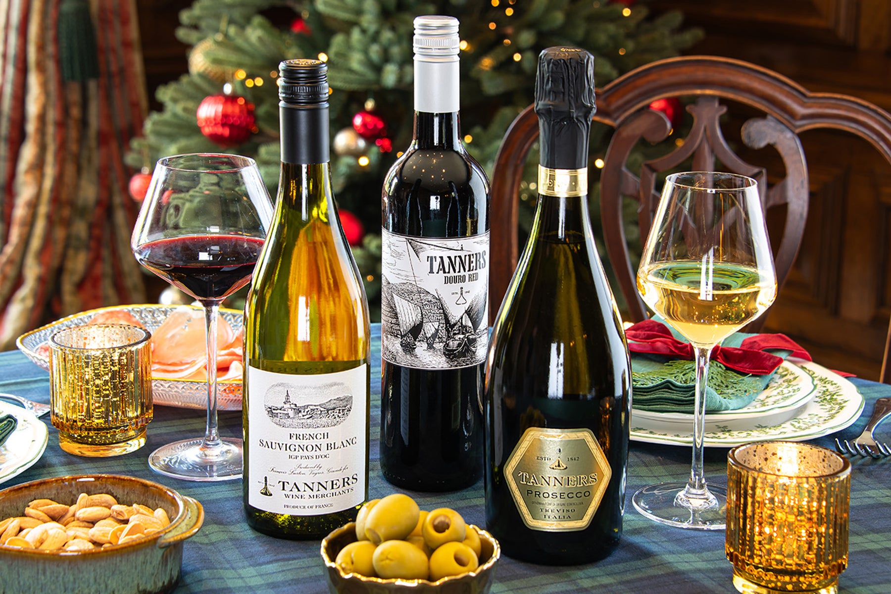 Three wine bottles including a French Sauvignon Blanc, Douro Red and Tanners Prosecco, alongside two wine glasses on a decorated dining table with a plaid tablecloth, candles, olives, and a Christmas tree in the background.