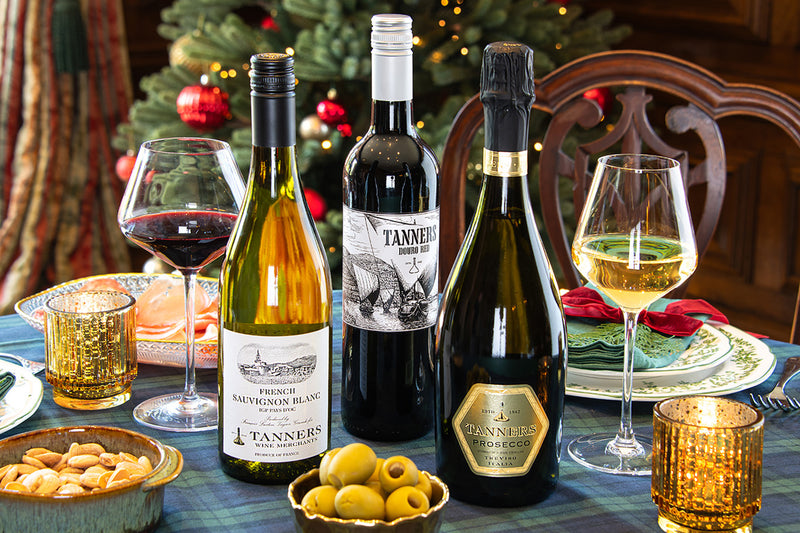 Three wine bottles including a French Sauvignon Blanc, Douro Red and Tanners Prosecco, alongside two wine glasses on a decorated dining table with a plaid tablecloth, candles, olives, and a Christmas tree in the background.