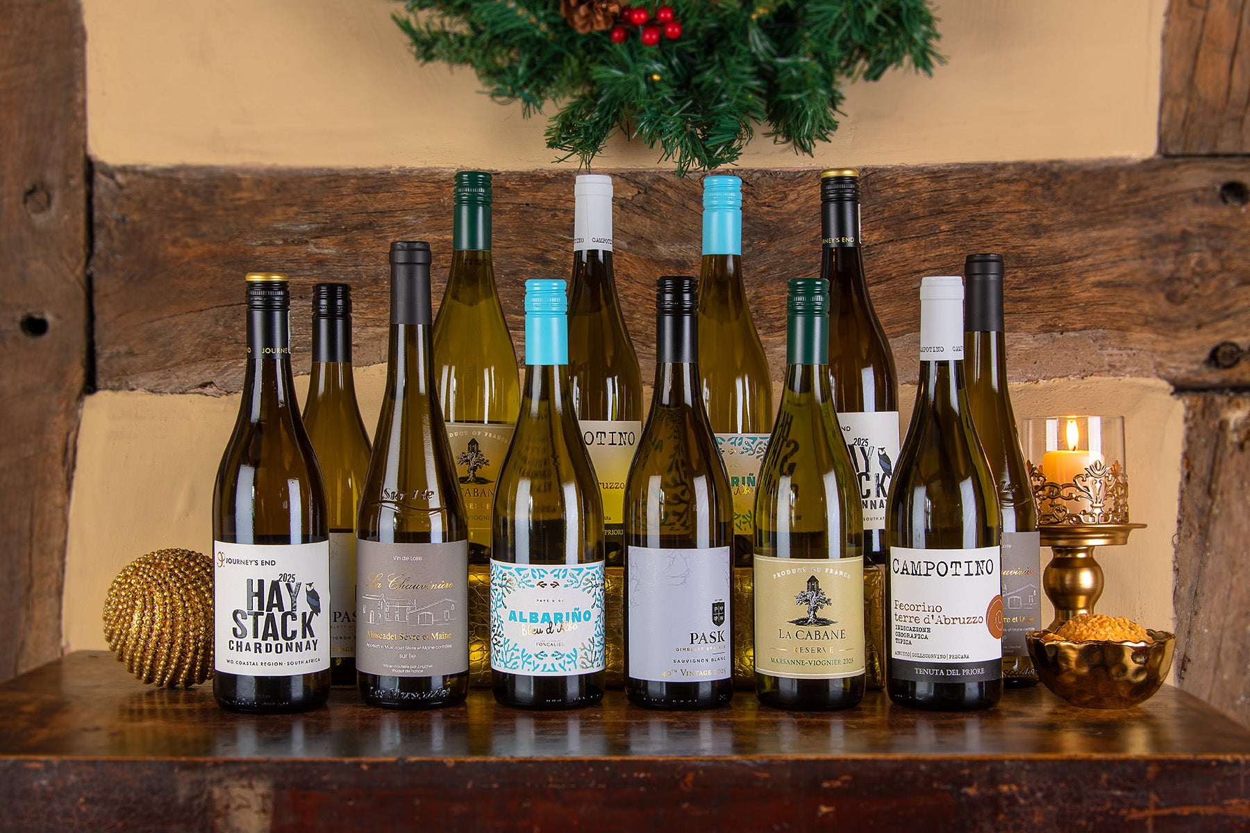 A group of wine bottles arranged on a wooden shelf against a rustic wood panel background, with decorative candles and greenery visible. Bottles include a Hay Stack Chardonnay, Bleu d'Alba Albariño, Pask Chardonnay, La Cabane, and a Campotino.