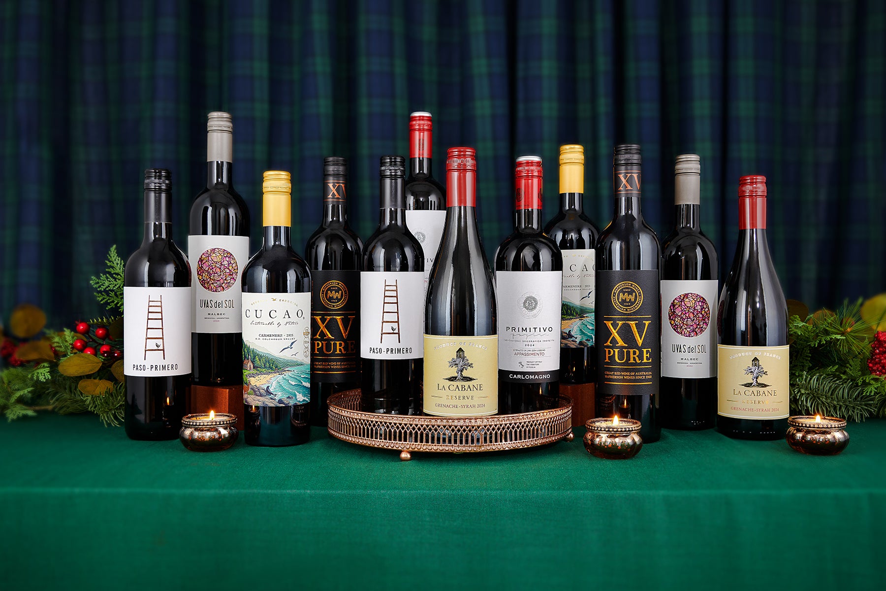 A group of wine bottles arranged on a green tablecloth with a green plaid backdrop. Several bottles including a Paso-Primero, UVAS del Sol, Cucao, XV Pur, La Cabane, and a Primitivo are displayed on and around a round metal tray, with small gold tea lights placed in front of them.