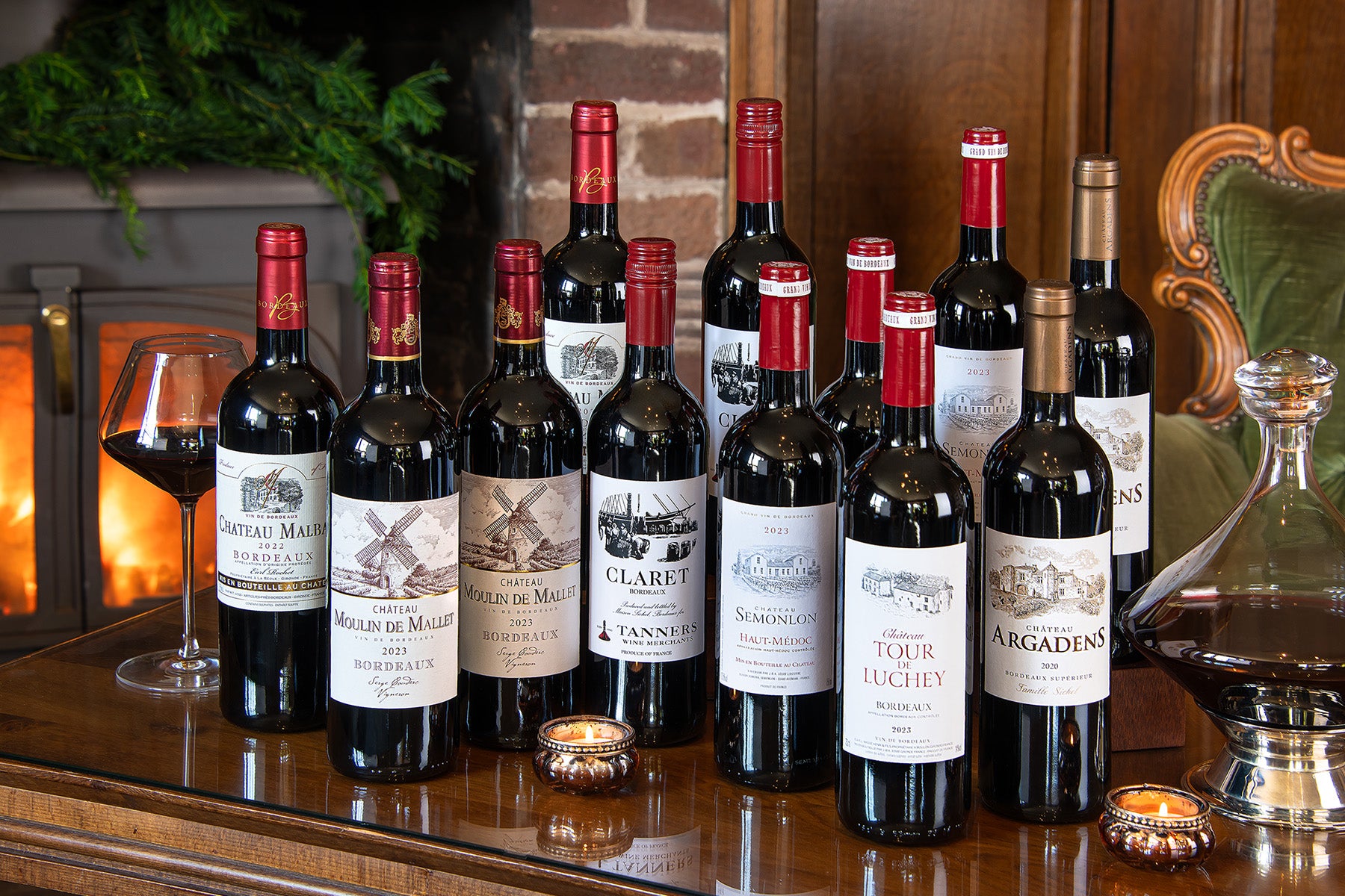A group of twelve wine bottles from renowned Bordeaux estates, including Chateau Malbat, Chateau Semonlon, and Chateau Argadens, arranged on a polished wooden table alongside a glass of red wine and candles.