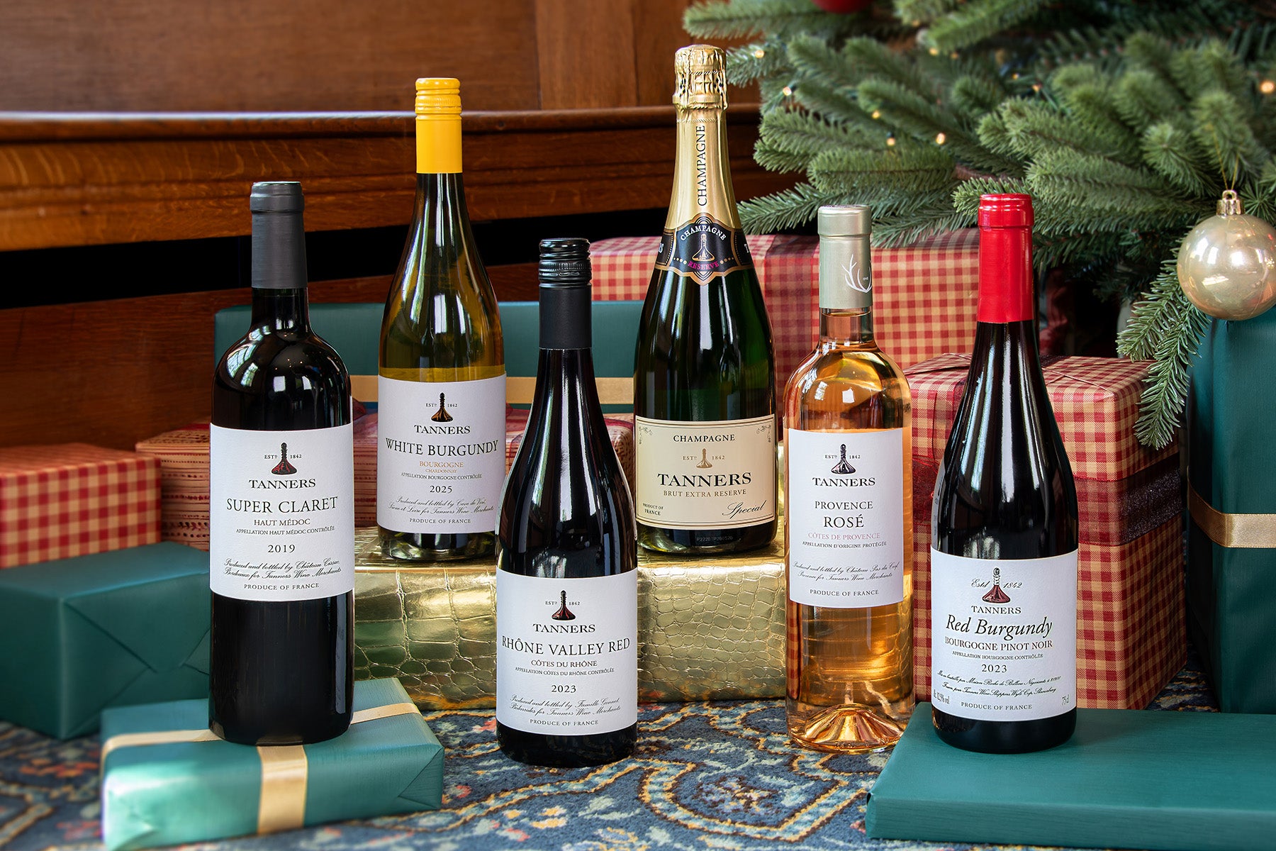 Six Tanners wine and sparkling wine bottles arranged on a reflective surface with wrapped gifts and a Christmas tree in the background. The bottles include a Super Claret, White Burgundy, Rhone Valley Red, Champagne Brut, Provence Rose, and a Red Burgundy.