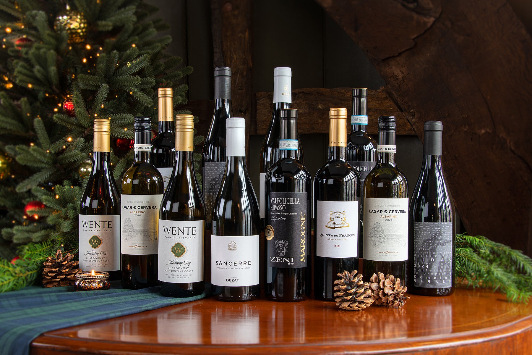 A group of wine bottles arranged on a wooden table with pinecones and a decorated Christmas tree in the background. Including a Wente Chardonnay, Lagar D Cervera, Sancerre, Marogne Zeni and a Domain Giraud.