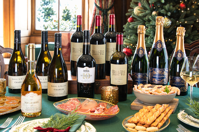 A festive holiday table display featuring an assortment of wine and sparkling wine bottles, including Bellevue, Noval 10 Year Old Tawny Port, André Clouet Grande Réserve, Brut Champagne, Chablis, and a  Tokaji Aszú 5 Puttonyos, Puklus Pincészet alongside plates of food, glasses, and a Christmas tree in the background.