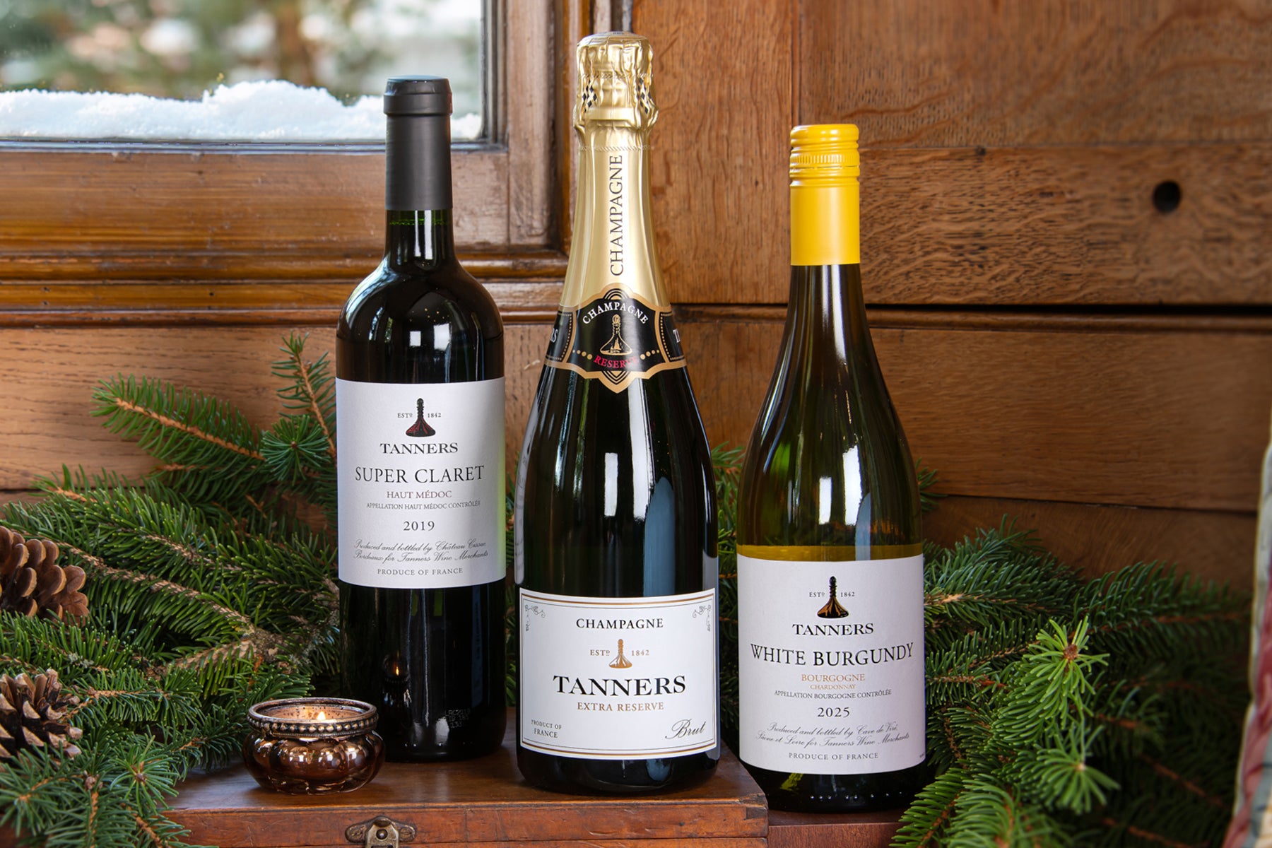 Three bottles of Tanners wine and champagne including a Super Claret, White Burgundy and Tanners Extra Reserve displayed on a wooden shelf with evergreen branches and a small candle, set against a rustic wood-paneled wall.