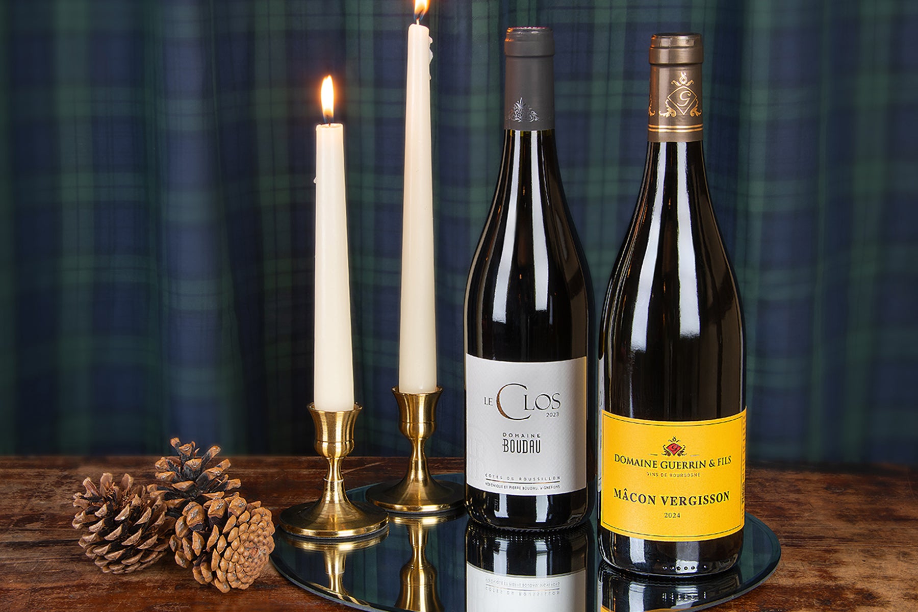 Two wine bottles including a Domaine Boudau and a Mâcon-Vergisson displayed on a round mirror tray alongside two lit white taper candles in brass holders and pinecones, set against a dark green plaid fabric backdrop on a wooden surface.