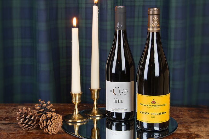 Two wine bottles including a Domaine Boudau and a Mâcon-Vergisson displayed on a round mirror tray alongside two lit white taper candles in brass holders and pinecones, set against a dark green plaid fabric backdrop on a wooden surface.
