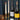 Two wine bottles including a Domaine Boudau and a Mâcon-Vergisson displayed on a round mirror tray alongside two lit white taper candles in brass holders and pinecones, set against a dark green plaid fabric backdrop on a wooden surface.