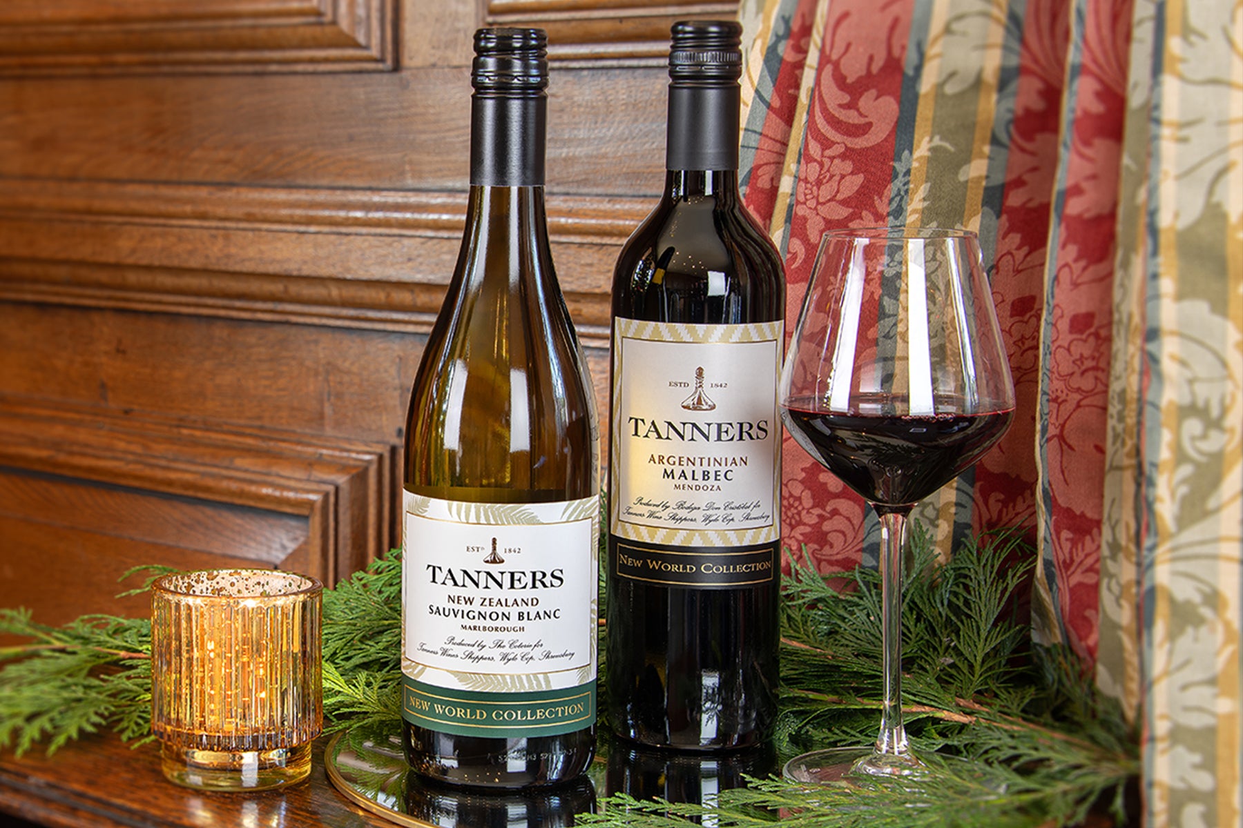 Two bottles of Tanners wine, including a New Zealand Sauvignon Blanc white wine bottle and an Argentinian Malbec red wine bottle, displayed on a gold tray with a filled wine glass, greenery, and a candle against a wooden paneled wall.