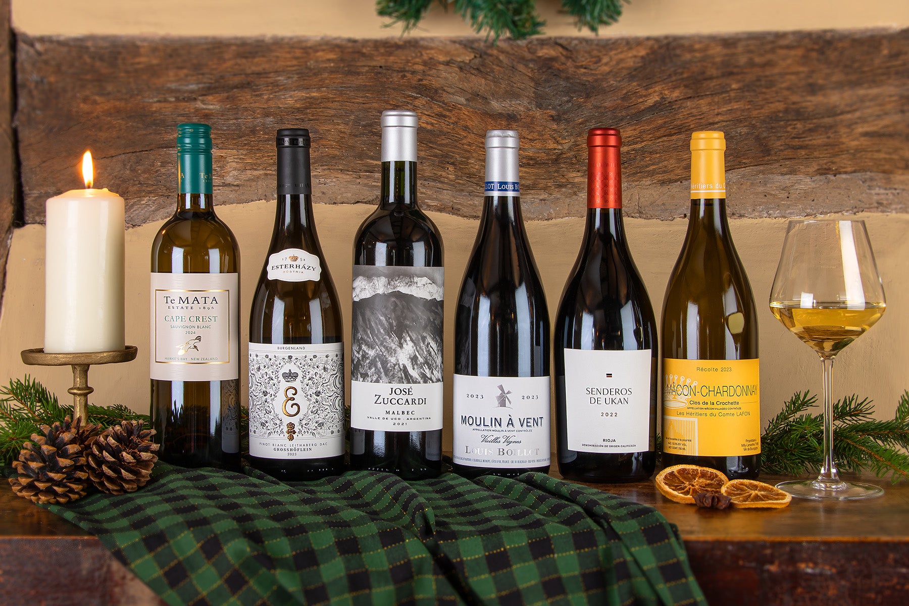 Six wine bottles including a Te Mata Cape Crest Sauvignon Blanc, Estoras Pinot Noir, José Zuccardi Malbec, Moulin-à-Vent, Senderos De Ukan and a Macon-Chardonnay alongside, a glass of white wine arranged on a rustic wooden table with a green plaid cloth, pinecones, a lit candle, dried orange slices, and holiday greenery in the background.