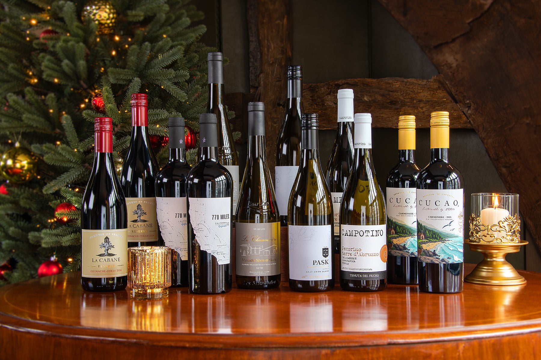 A group of assorted wine bottles arranged on a wooden table in front of a decorated Christmas tree and rustic wooden wall. Notable brands include La Cabane, 770 Miles, PASK, Cucao and Campotino.