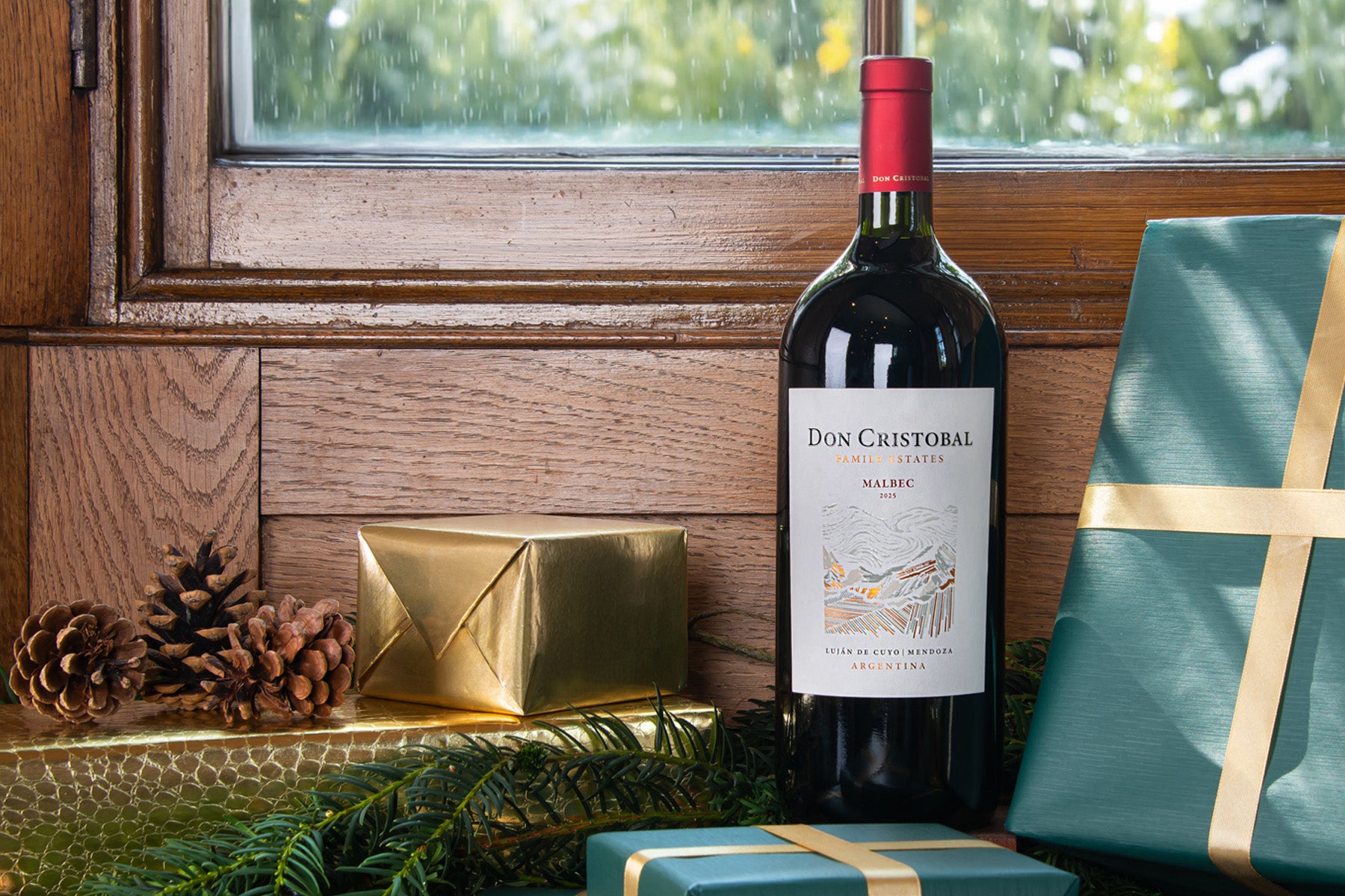 A bottle of Don Cristobal Malbec wine stands on a wooden windowsill decorated with pine branches, pinecones, and wrapped gift boxes in teal and gold.