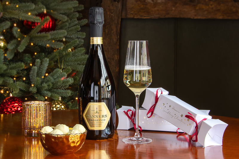 Bottle of Tanners Prosecco and Truffles with glass on a wooden table in front of a Christmas tree with festive tea light and some truffles in a golden bowl.