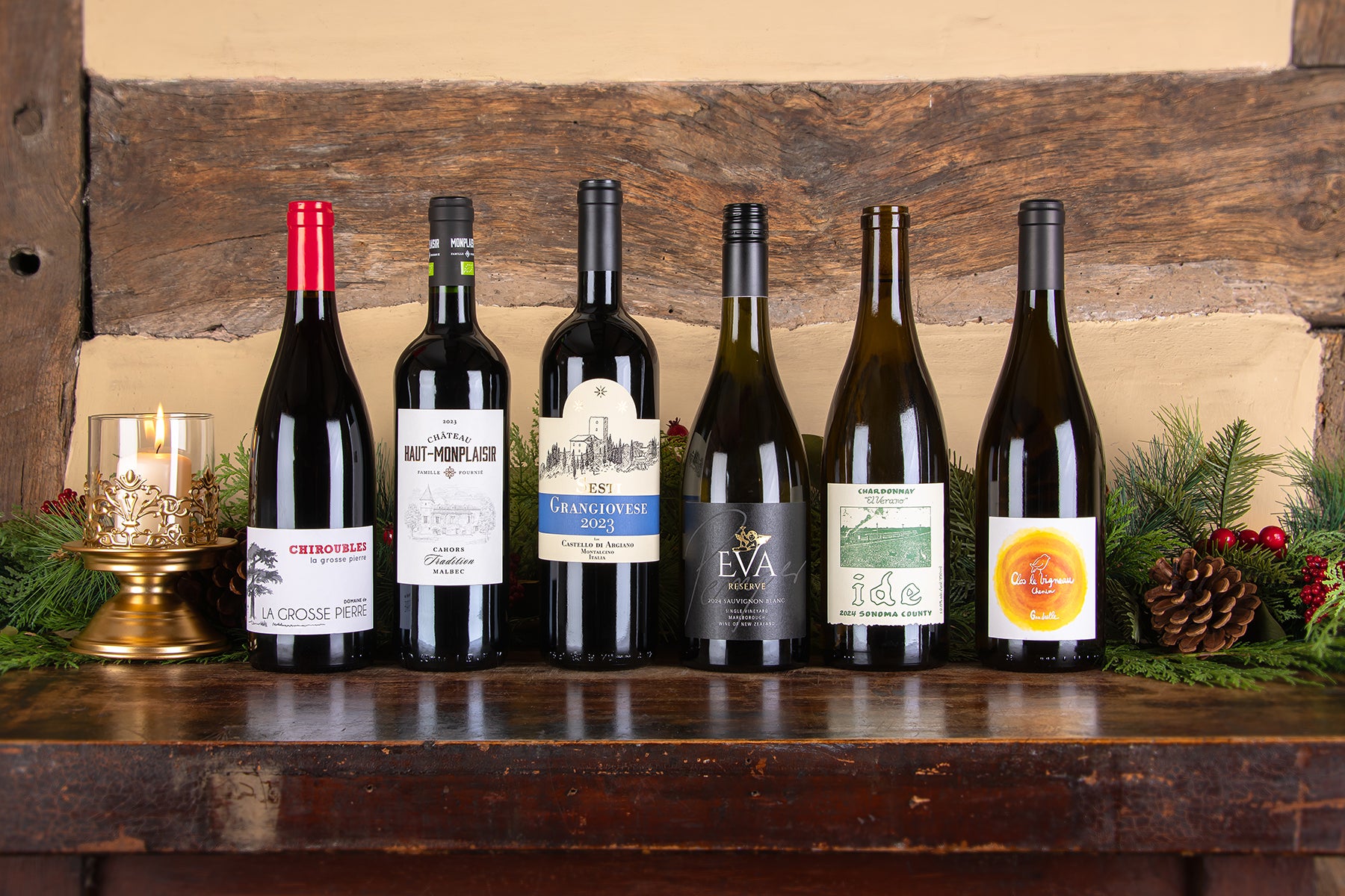 Six wine bottles including favourites  Chiroubles, Cahors,Tradition, Malbec, Grangiovese, Eva, Ide and Vouvray lined up on a rustic wooden shelf with a candle holder, evergreen branches, pinecones, and a beige wood-paneled background.