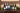 Six wine bottles including favourites  Chiroubles, Cahors,Tradition, Malbec, Grangiovese, Eva, Ide and Vouvray lined up on a rustic wooden shelf with a candle holder, evergreen branches, pinecones, and a beige wood-paneled background.
