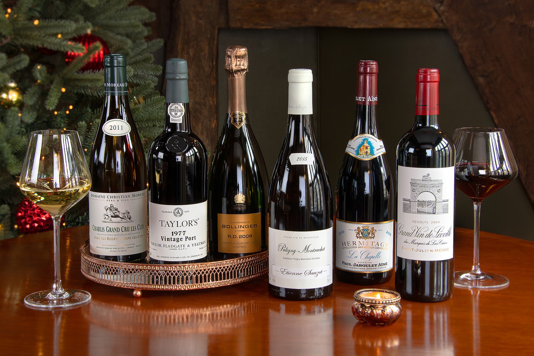 Six wine bottles arranged on a wooden table with wine glasses, a candle, and a decorated Christmas tree in the background. Bottles include a Chablis Grand Cru Les Clos, Taylor's 1977 Vintage Port, Bollinger RD, Extra Brut Champagne, Puligny-Montrachet, Hermitage Blanc, La Chapelle and a Château Léoville.