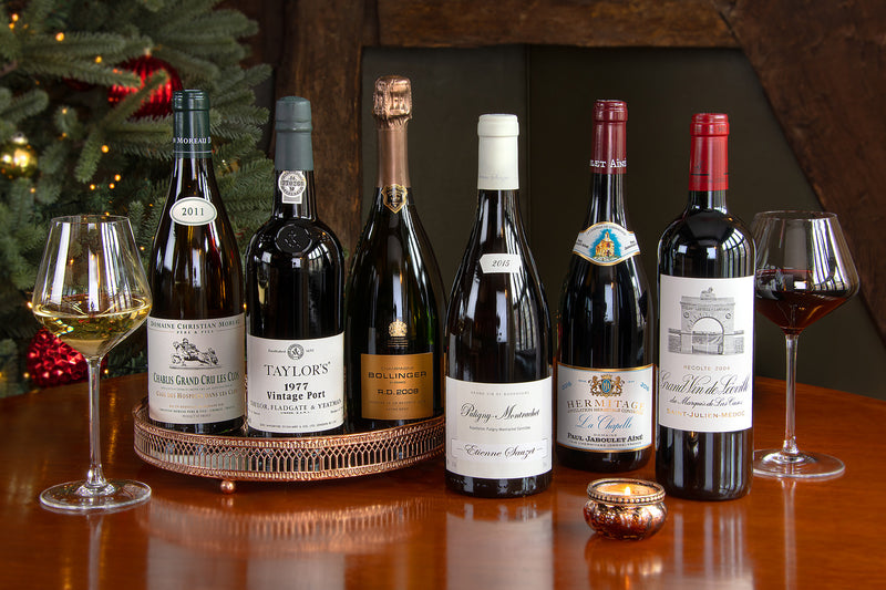 Six wine bottles arranged on a wooden table with wine glasses, a candle, and a decorated Christmas tree in the background. Bottles include a Chablis Grand Cru Les Clos, Taylor's 1977 Vintage Port, Bollinger RD, Extra Brut Champagne, Puligny-Montrachet, Hermitage Blanc, La Chapelle and a Château Léoville.