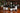 Six wine bottles arranged on a wooden table with wine glasses, a candle, and a decorated Christmas tree in the background. Bottles include a Chablis Grand Cru Les Clos, Taylor's 1977 Vintage Port, Bollinger RD, Extra Brut Champagne, Puligny-Montrachet, Hermitage Blanc, La Chapelle and a Château Léoville.