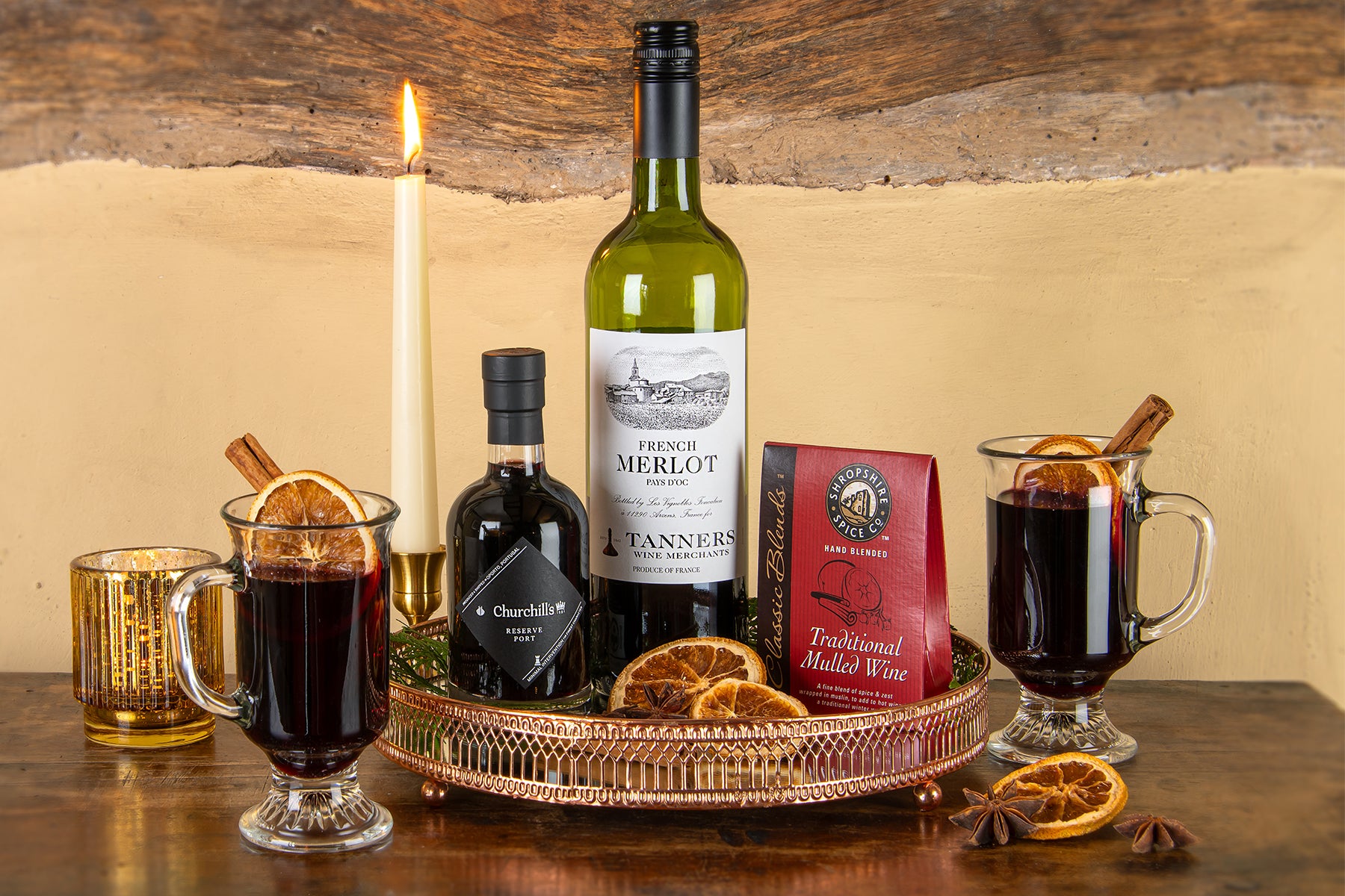 Tanners Marvellous Mulled Wine Kit on a wooden table featuring a bottle of Tanners French Merlot wine, a bottle of Churchill's Reserve Port, mulled wine spices including orange slices and cinnamon sticks, arranged around a copper tray