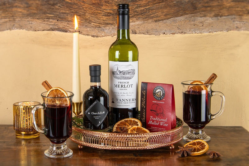 Tanners Marvellous Mulled Wine Kit on a wooden table featuring a bottle of Tanners French Merlot wine, a bottle of Churchill's Reserve Port, mulled wine spices including orange slices and cinnamon sticks, arranged around a copper tray