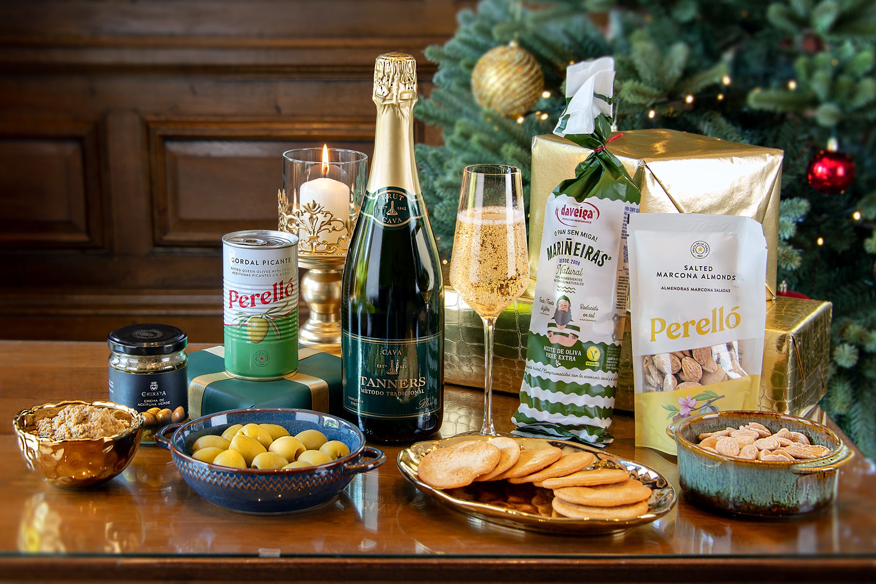 A Taste of Tanners at Home Christmas Gift featuring a bottle of Tanners Cava, Perello salted almonds, green olives, biscuits, and tapenade in front of a decorated Christmas tree