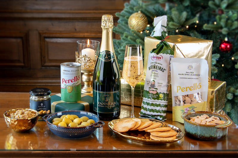 A Taste of Tanners at Home Christmas Gift featuring a bottle of Tanners Cava, Perello salted almonds, green olives, biscuits, and tapenade in front of a decorated Christmas tree