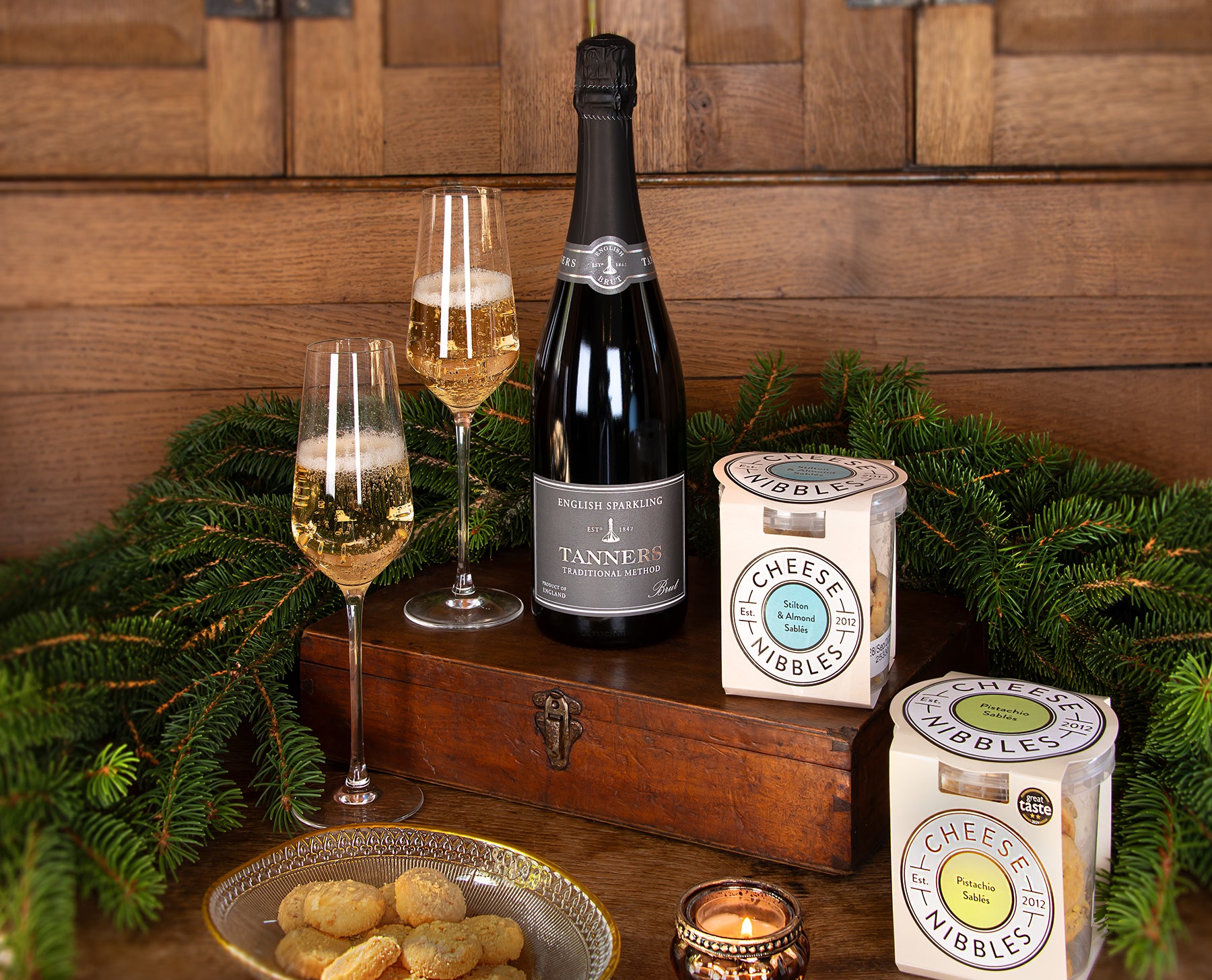 A styled holiday gift set featuring a bottle of Tanners English Sparkling Wine, two filled champagne flutes, two boxes of Cheese Nibbles, and a plate of cookies arranged on a wooden chest with evergreen branches and a candle against a rustic wood-paneled background.