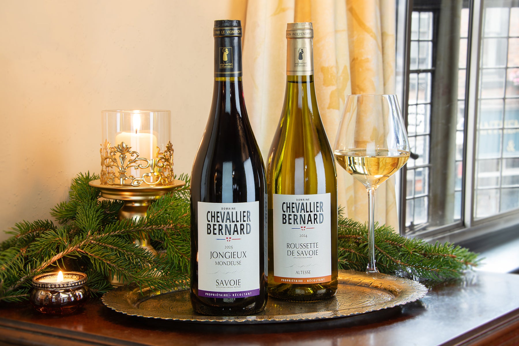 Two bottles of Chevallier Bernard wine and a filled wine glass on a decorative tray, styled with greenery and candles near a window.