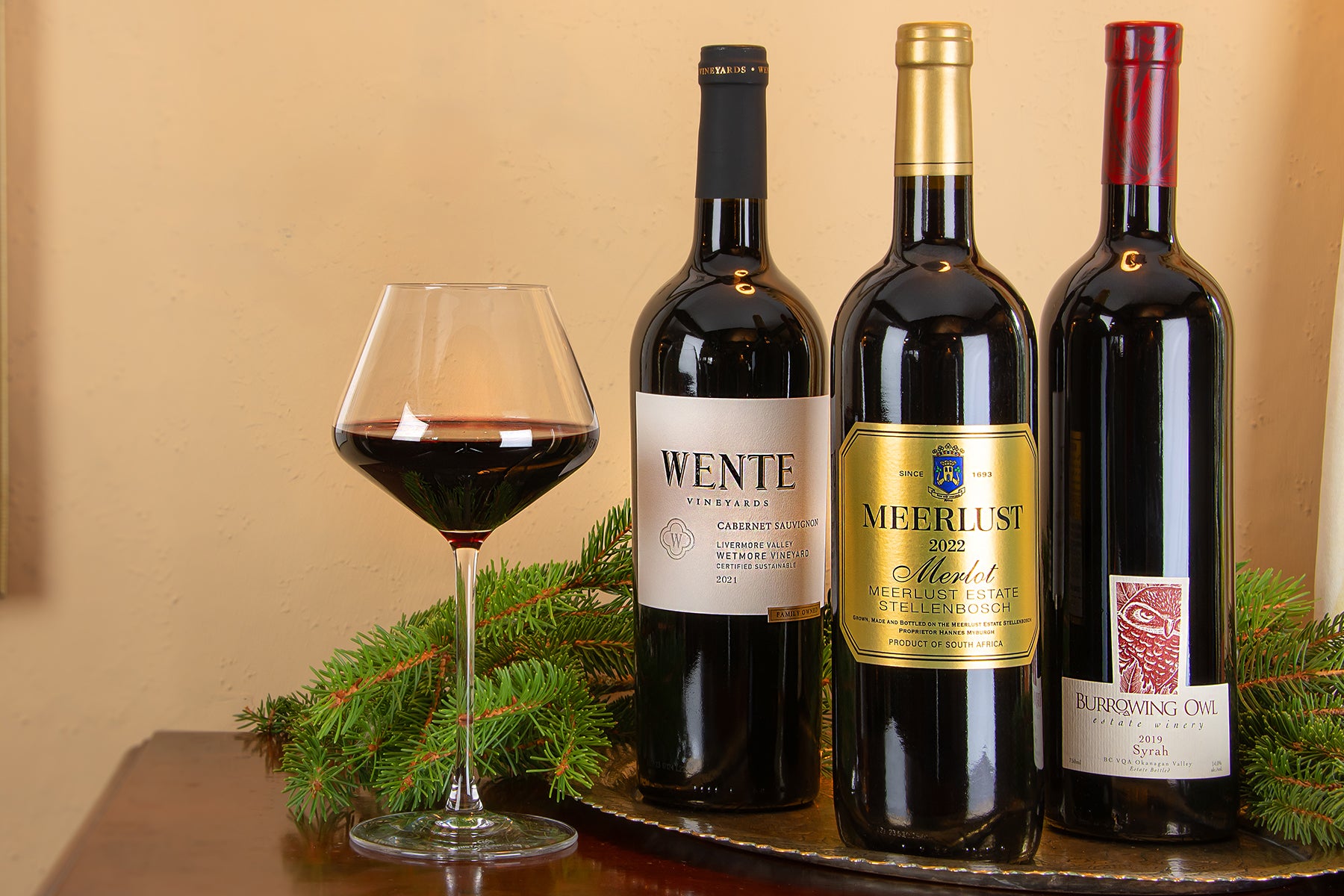 Three bottles of red wine and a filled wine glass arranged on a tray with evergreen branches against a warm beige wall. Includes a Wente 'Charles Wetmore' Cabernet Sauvignon, Meerlust Merlot, and a Burrowing Owl Syrah.