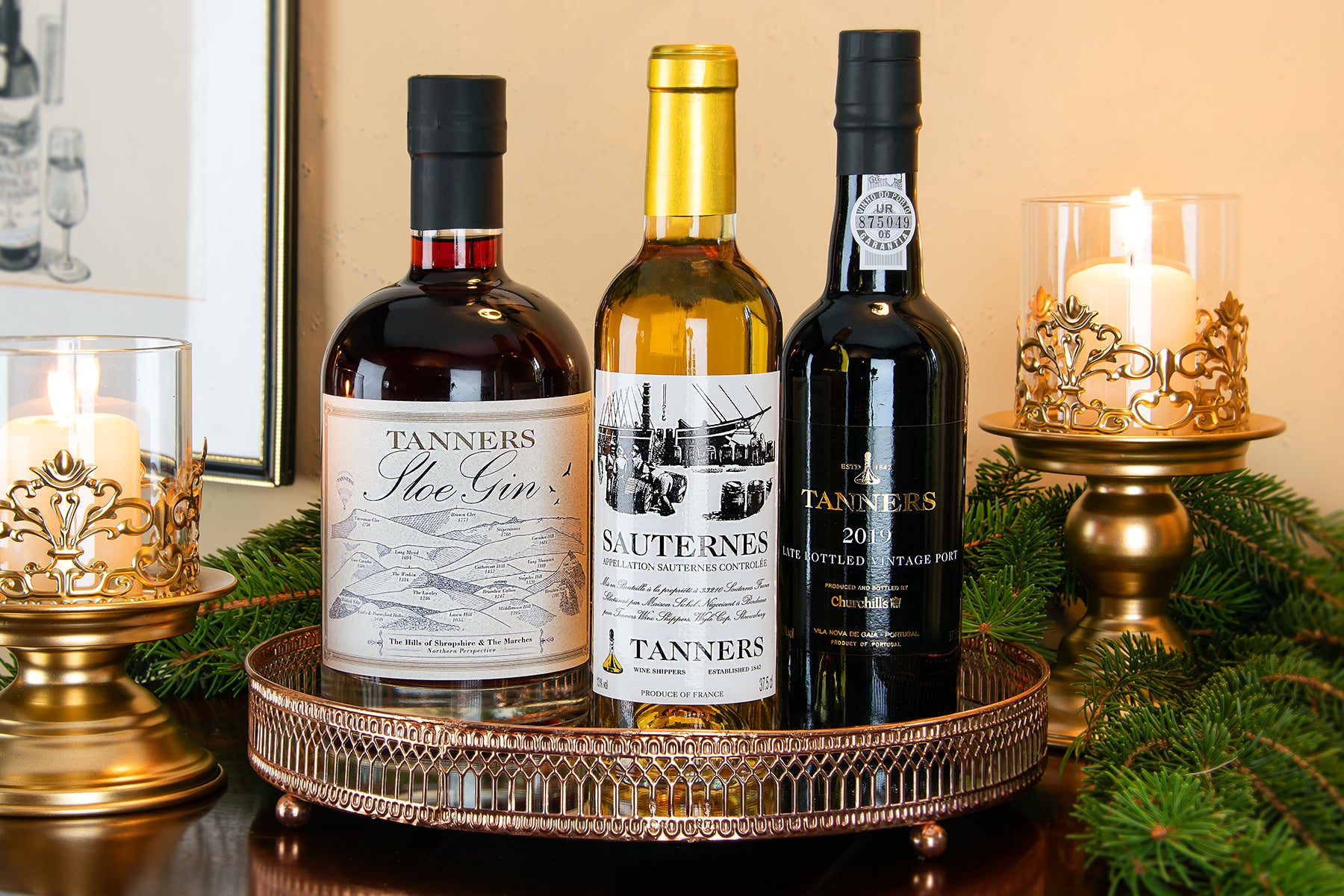 Three bottles of Tanners spirits including a Sloe Gin, Sauternes and Vintage Port arranged on a decorative copper tray with lit candles and greenery in the background.