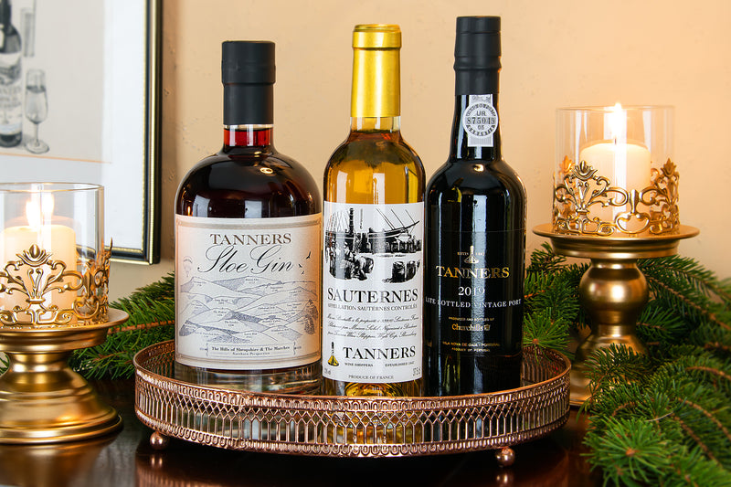 Three bottles of Tanners spirits including a Sloe Gin, Sauternes and Vintage Port arranged on a decorative copper tray with lit candles and greenery in the background.