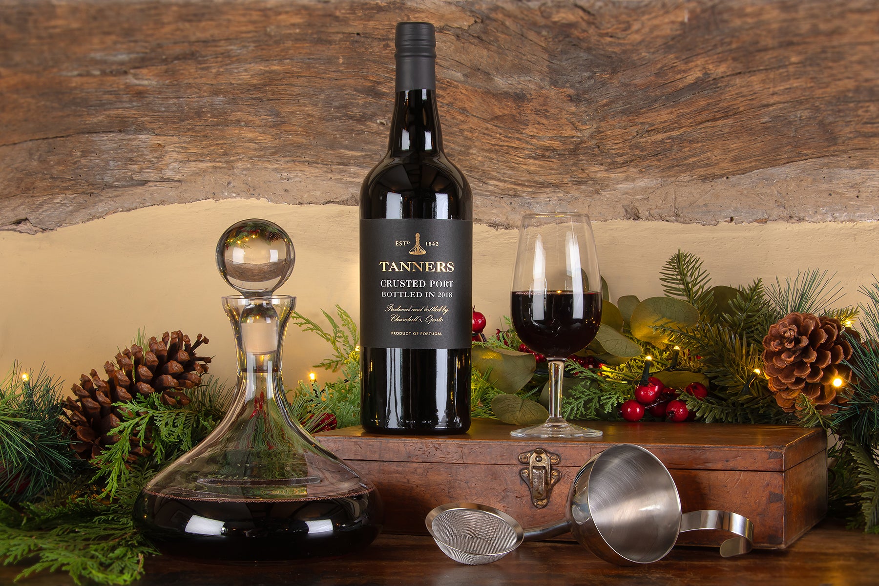 A bottle of Tanners Crusted Port stands on a wooden surface beside a filled wine glass, a clear glass decanter, metal pouring tools, and holiday greenery with pinecones and red berries against a rustic wood wall.