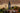 A bottle of Tanners Crusted Port stands on a wooden surface beside a filled wine glass, a clear glass decanter, metal pouring tools, and holiday greenery with pinecones and red berries against a rustic wood wall.