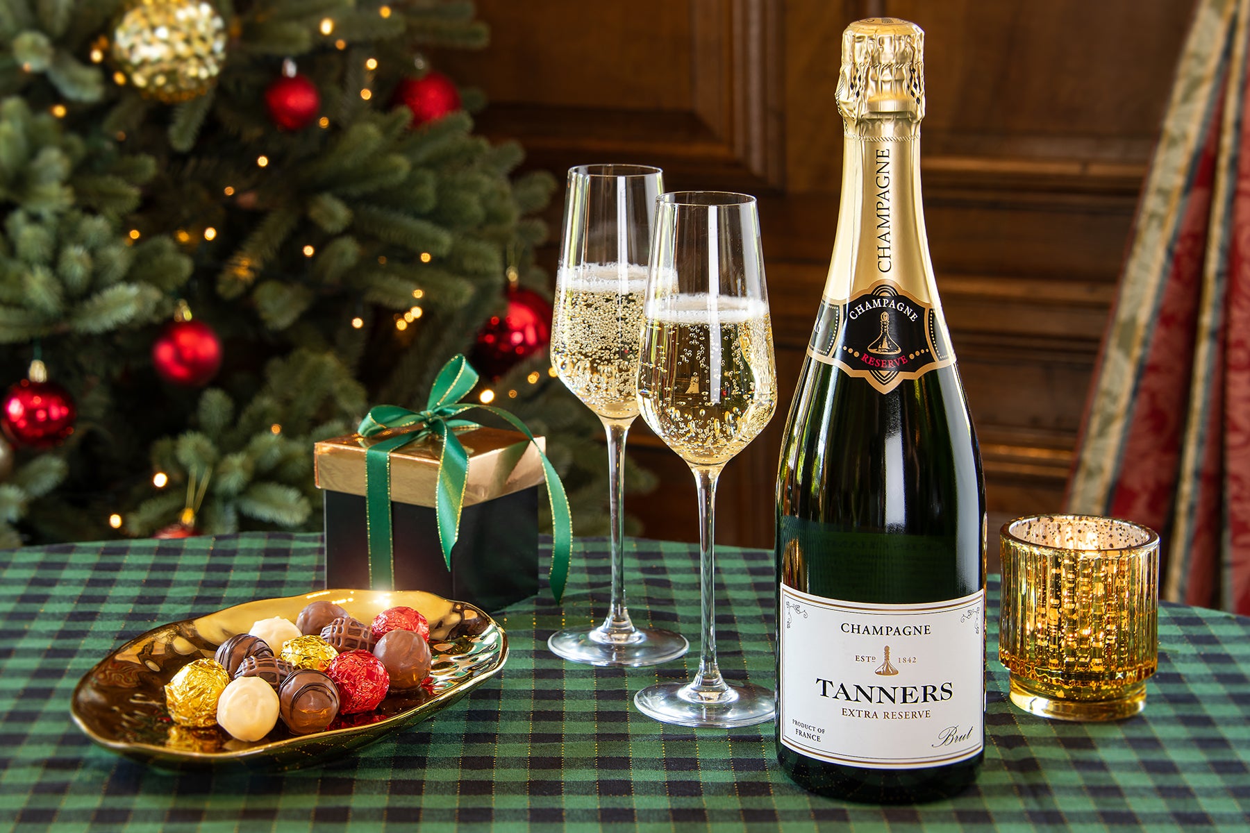 A bottle of Tanners Champagne Extra Reserve stands on a green plaid tablecloth beside two filled champagne flutes, a gold candle holder, a plate of assorted chocolates, and a wrapped gift box, with a decorated Christmas tree in the background.